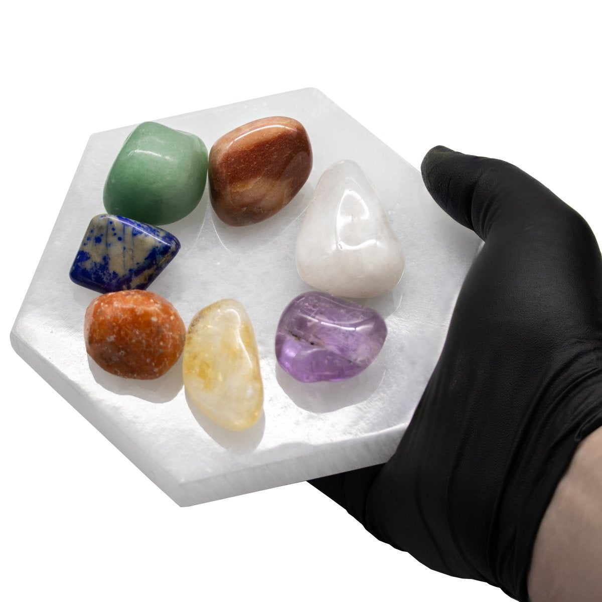 Seven colorful tumbled gemstones arranged on a white selenite hexagon plate held in a black-gloved hand.