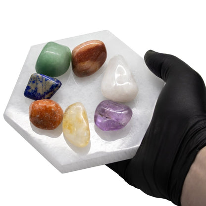 Seven colorful tumbled gemstones arranged on a white selenite hexagon plate held in a black-gloved hand.